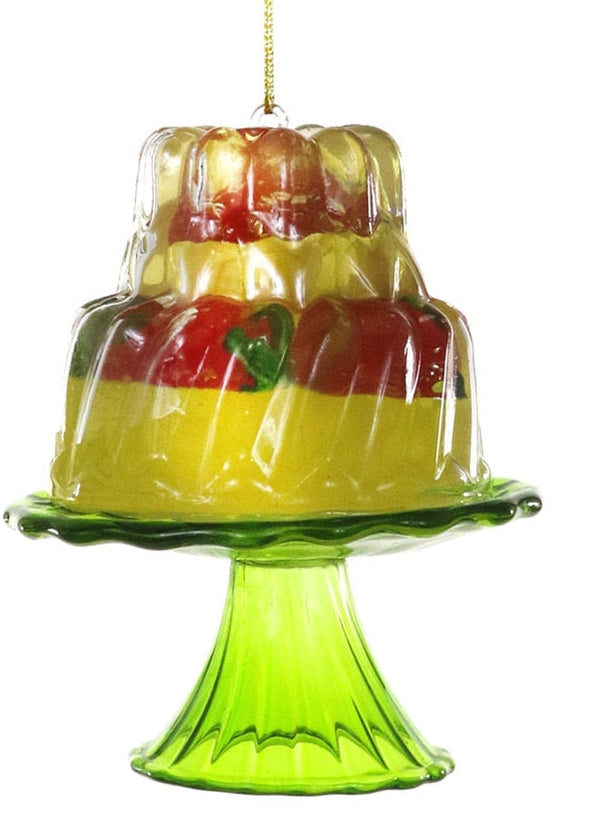 A decorative glass hanging ornament shaped like a two-tiered cake on a pedestal stand, with yellow and red layers and a green base, suspended by a gold string against a white background.