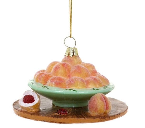 A hanging ornament shaped like a bowl of peaches on a wooden board, with a small pastry and a peach beside it, photographed against a plain white background.