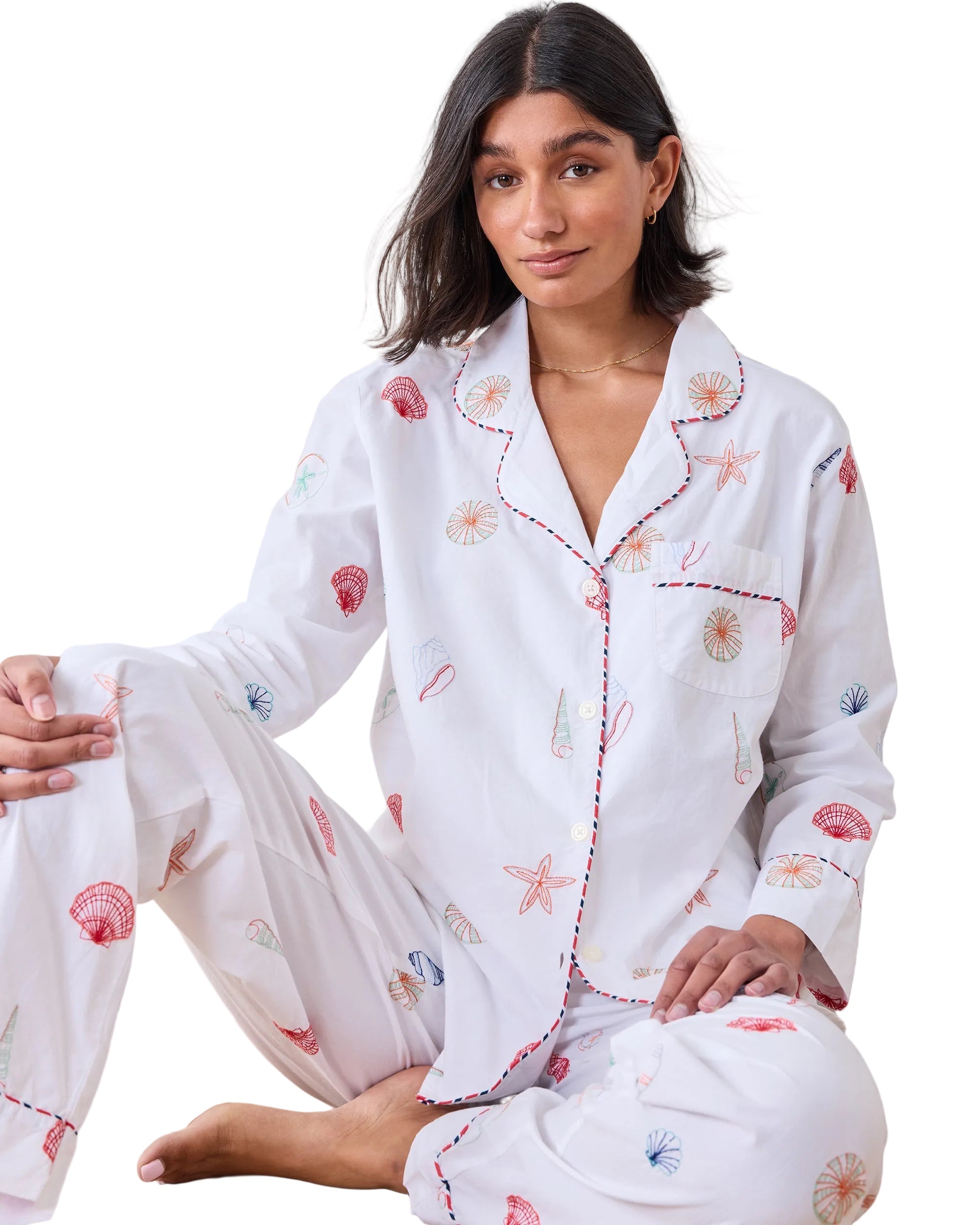 Woman wearing a white pajama set with floral embroidery sitting on a wooden floor.