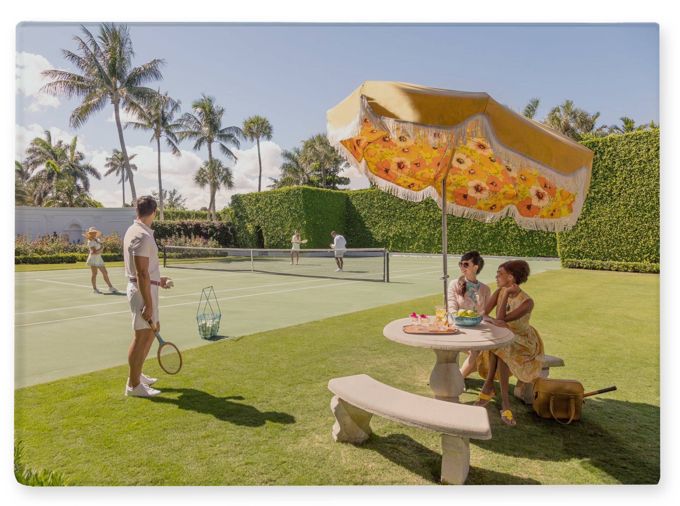 People playing tennis and sitting at a table with a floral umbrella in a park-like setting.