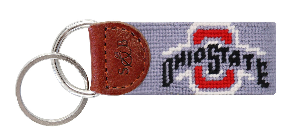 A needlepoint keychain featuring the Ohio State Buckeyes logo in red, white, and black on a lavender needlepoint canvas, attached to a brown leather tab with an embossed 'S&E' monogram and split metal key rings.