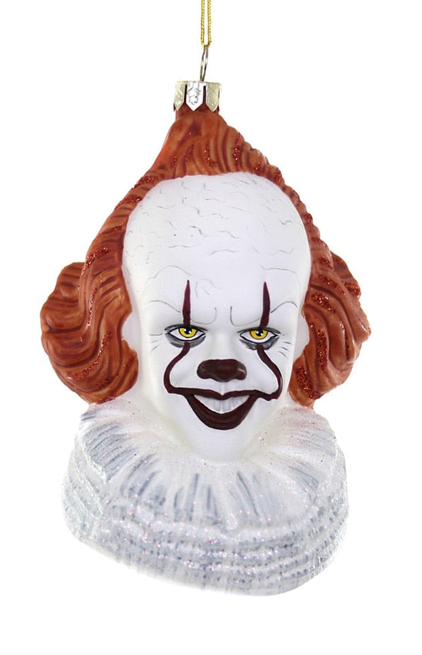 A Pennywise the Clown Christmas ornament featuring a white face with red and black painted makeup, brown hair, and a white ruffled collar, hanging from a gold string against a white background.