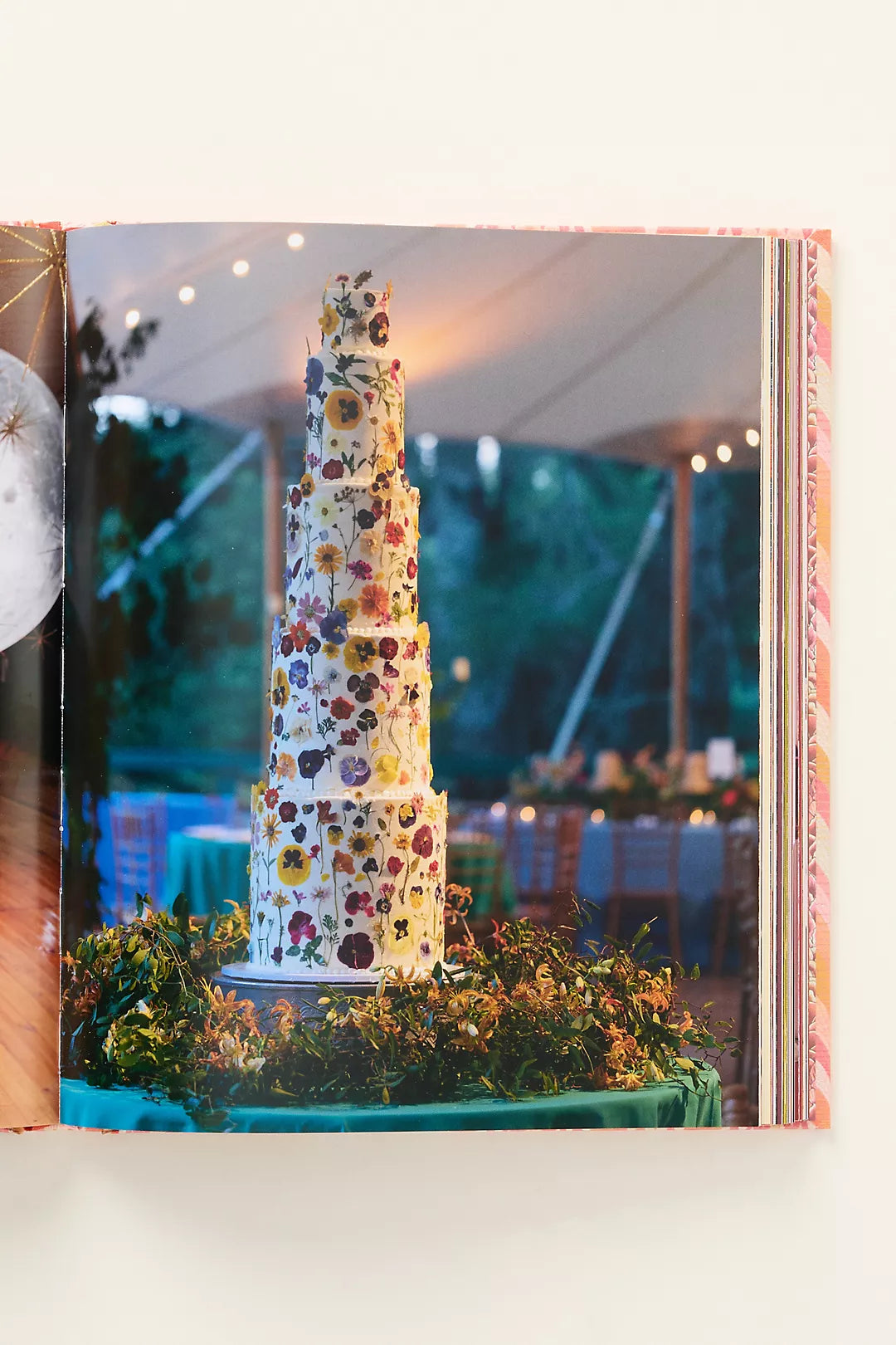 Floral tiered cake at an outdoor event with greenery and tables in the background.