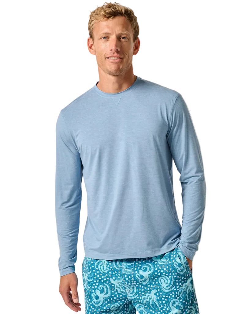 A man wearing a long-sleeved blue sunshirt and patterned swim shorts.