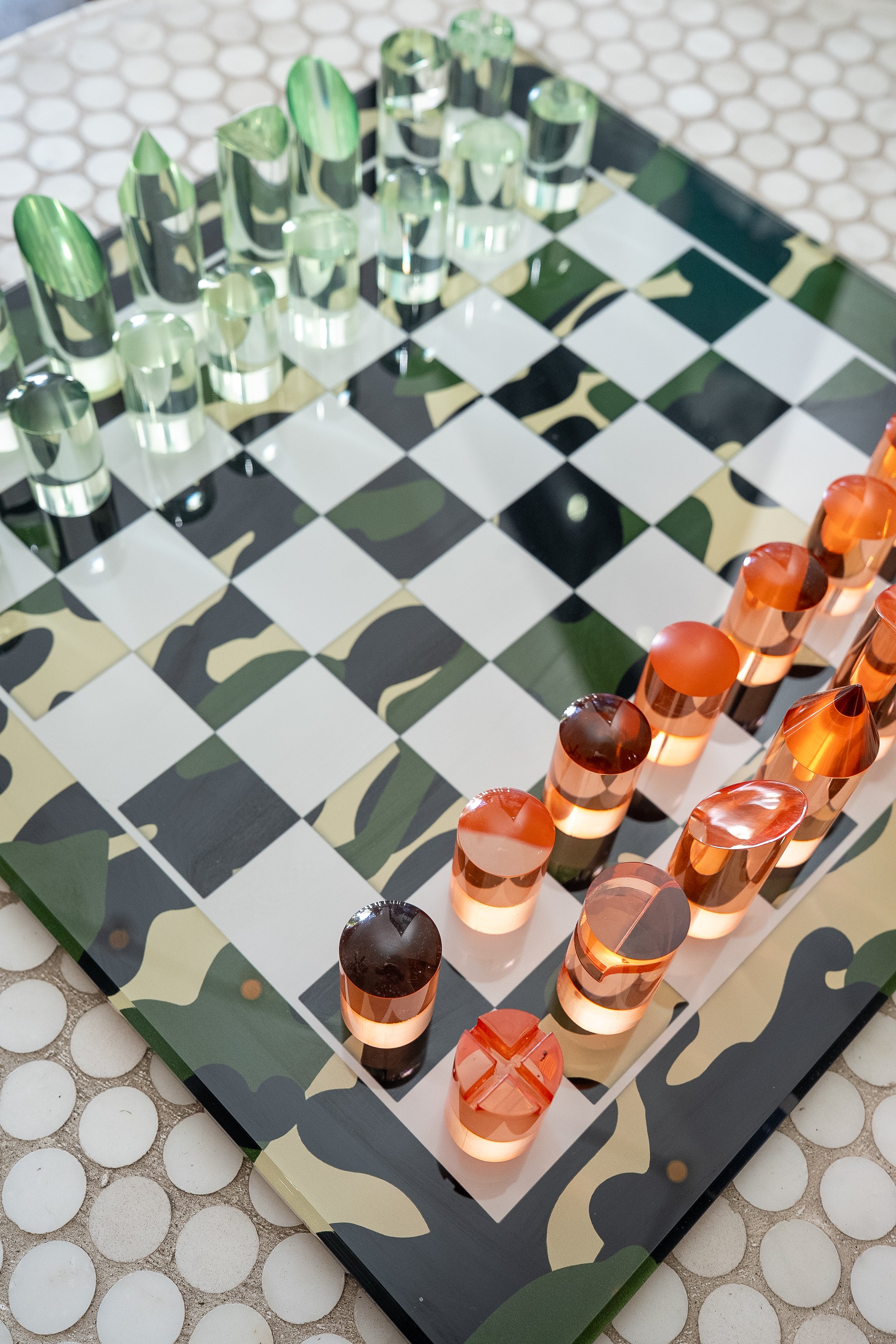 Camouflage-patterned chessboard with green, orange, and brown pieces on a checkered surface.
