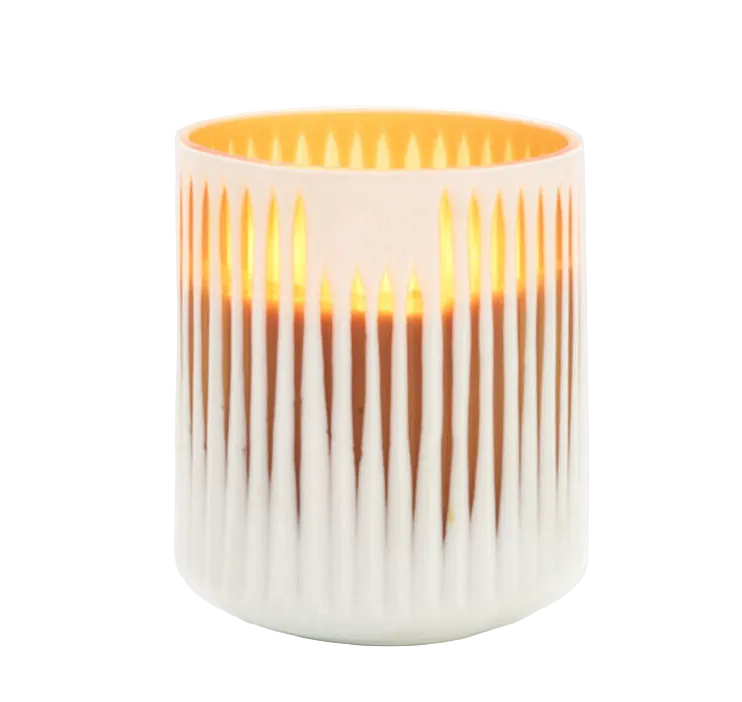A white glass candle container with vertical stripes, partially filled with a white substance, with a lit candle inside giving off a warm glow.