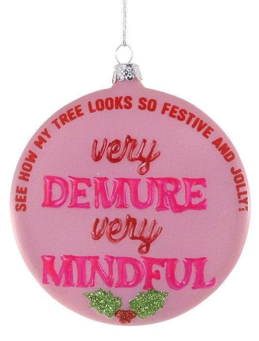 A round pink Christmas ornament with a silver hanging loop and silver cap, featuring the text 'very DEMURE very MINDFUL' in pink and red lettering, with green holly accents at the bottom, on a white background.