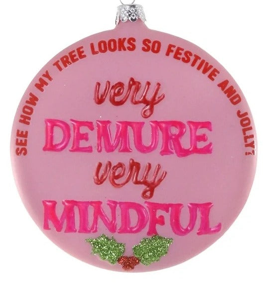 A round pink Christmas ornament with a silver hanging loop and silver cap, featuring the text 'very DEMURE very MINDFUL' in pink and red lettering, with green holly accents at the bottom, on a white background.