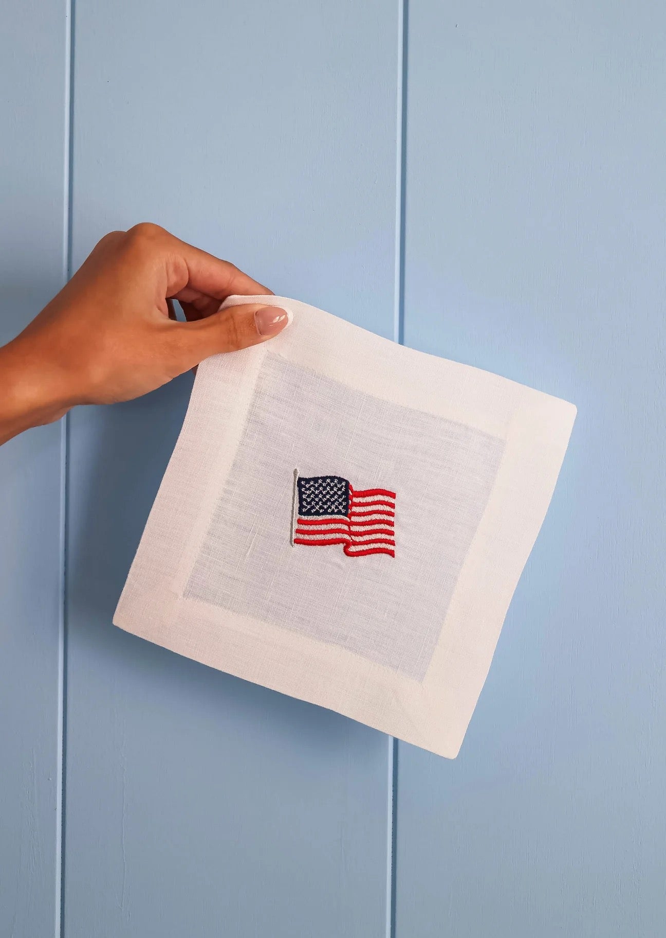 Hand holding a white napkin with an embroidered American flag design against a light blue background