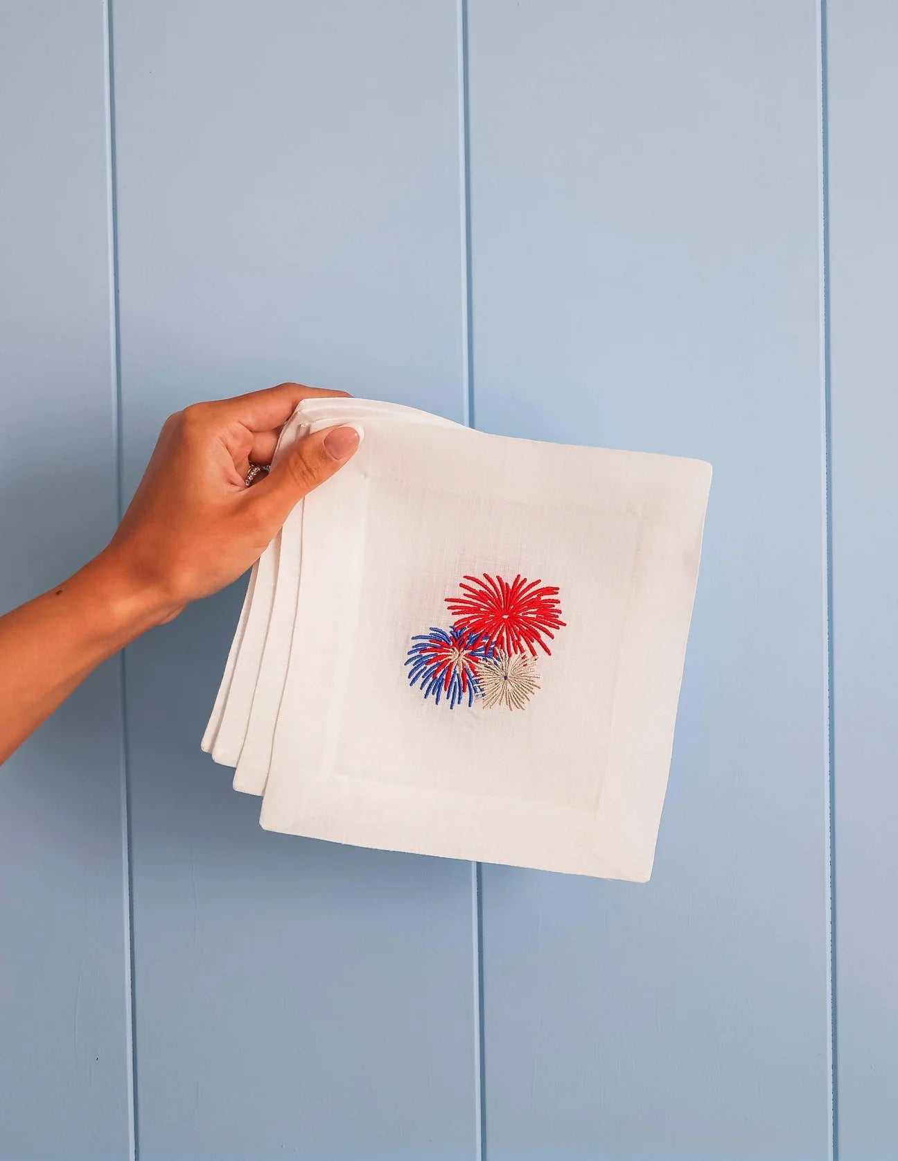 Hand holding a stack of white napkins with red, blue, and yellow firework designs against a light blue wooden panel background.