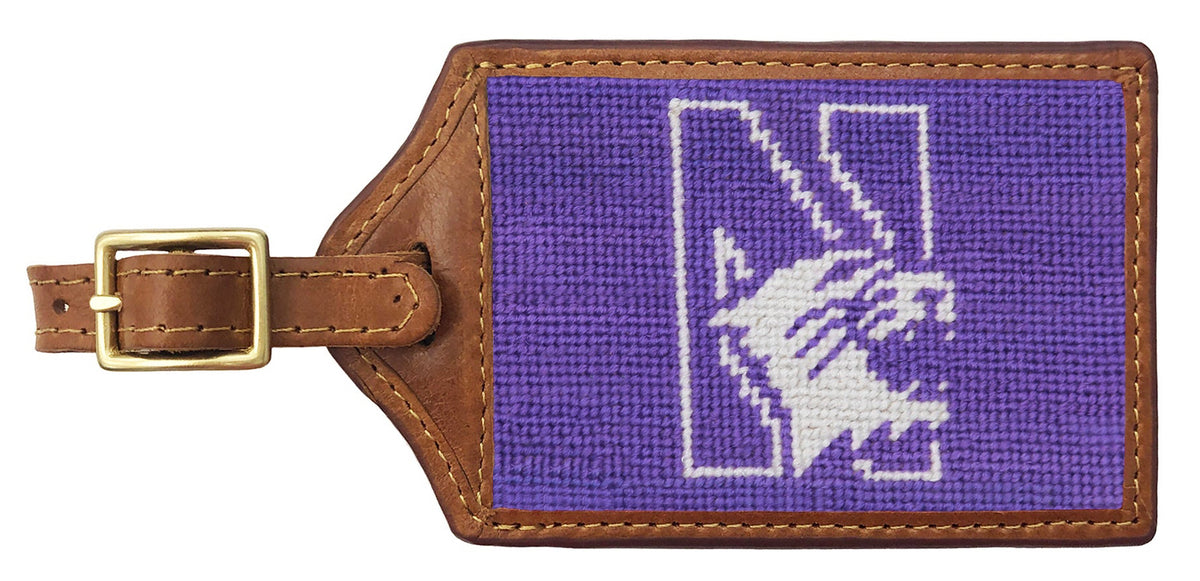 A rectangular brown leather luggage tag with a gold-tone buckle, featuring a purple woven canvas face with a white stylized logo.