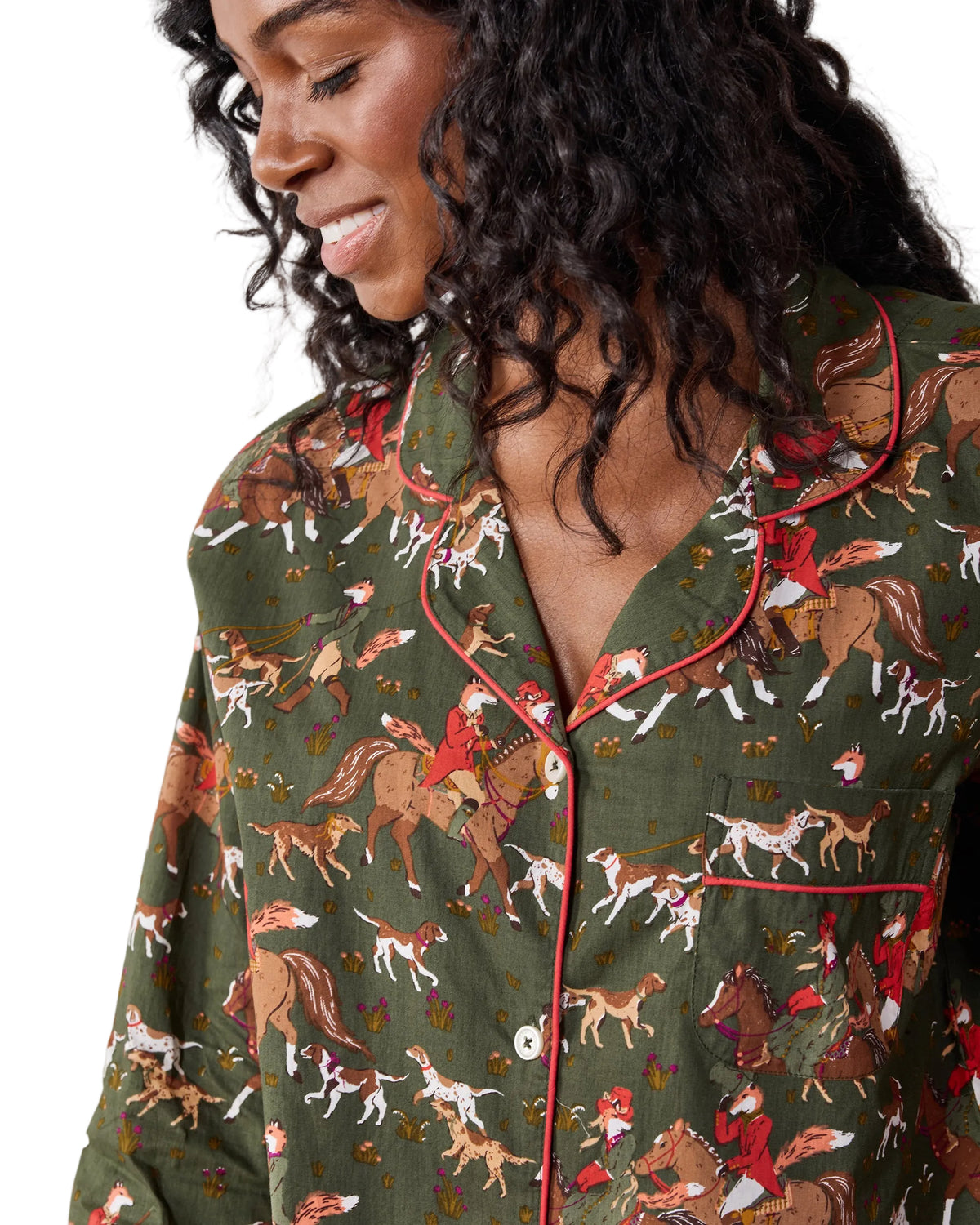 A close-up of a woman wearing an olive green pajama shirt with a detailed print of horses and riders in red, brown, and white, featuring red piping along the collar and placket.