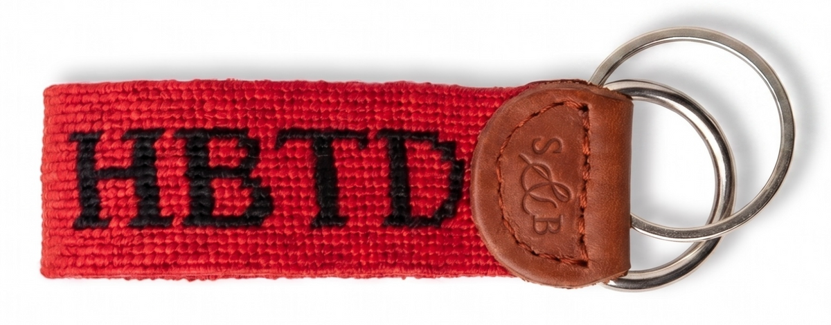 A red woven keychain strap with black embroidered letters reading 'HBTD' and a brown leather patch attached to two silver metal key rings, shown on a white background.