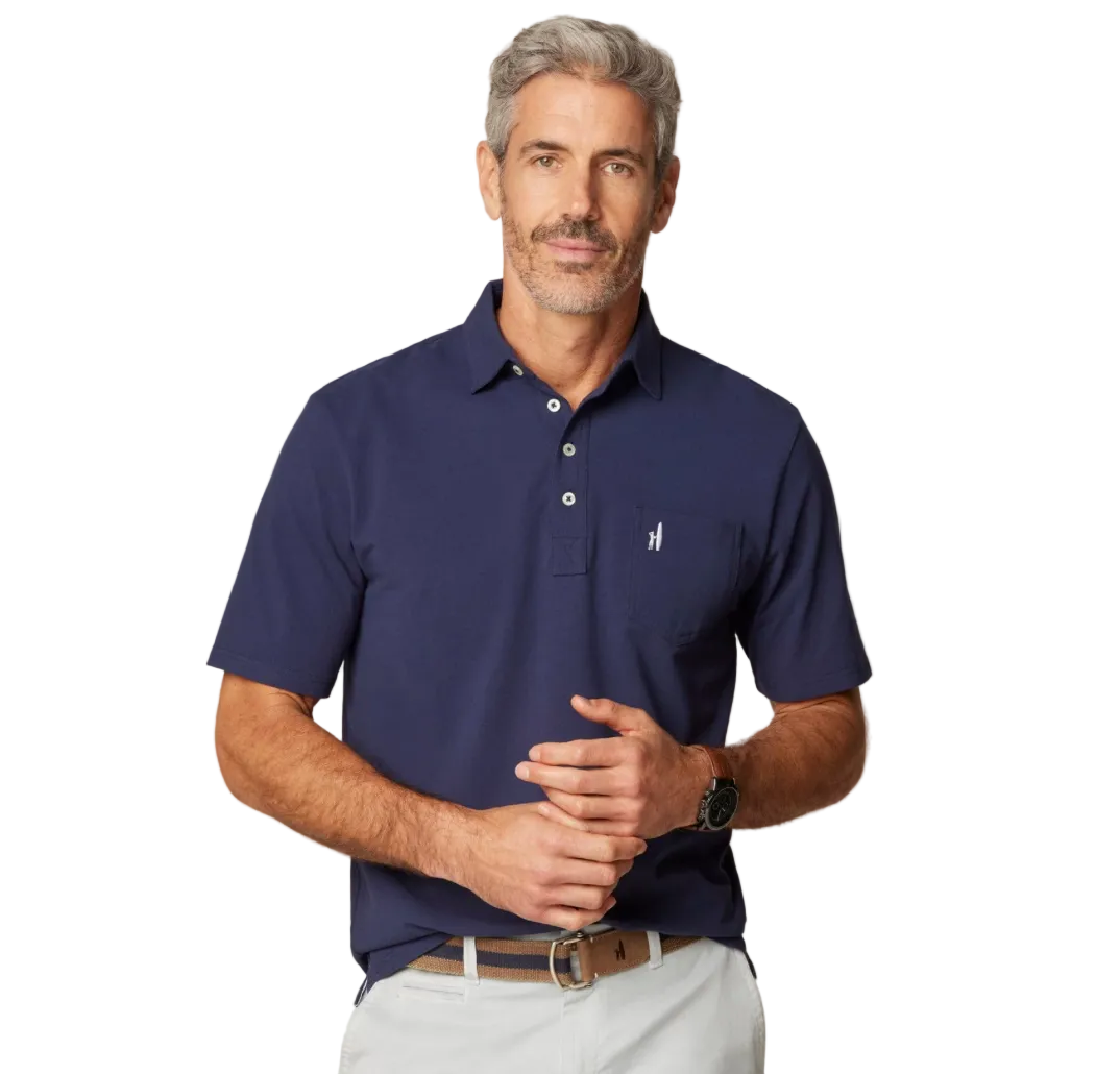 A man wearing a navy blue polo shirt with a collar, button-up neck, and short sleeves. He has a beard and is wearing a white pair of pants.