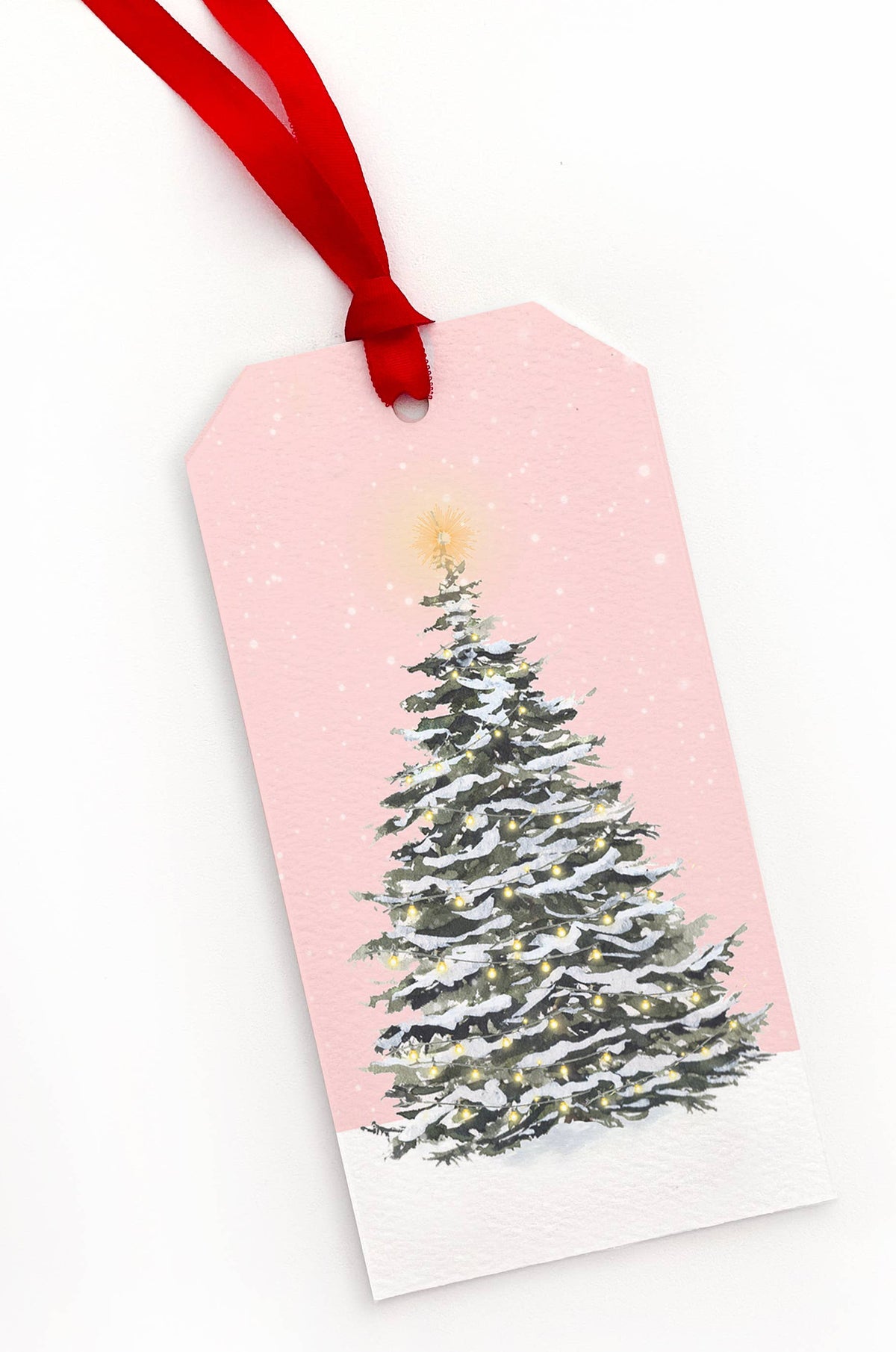 A rectangular gift tag with a red hanging ribbon, featuring a printed illustration of a snowy Christmas tree on a light pink background with a white bottom section.