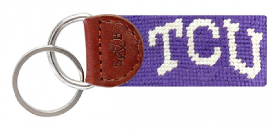 A purple needlepoint key fob with the text 'TCU' woven on it, attached to a brown leather fob.