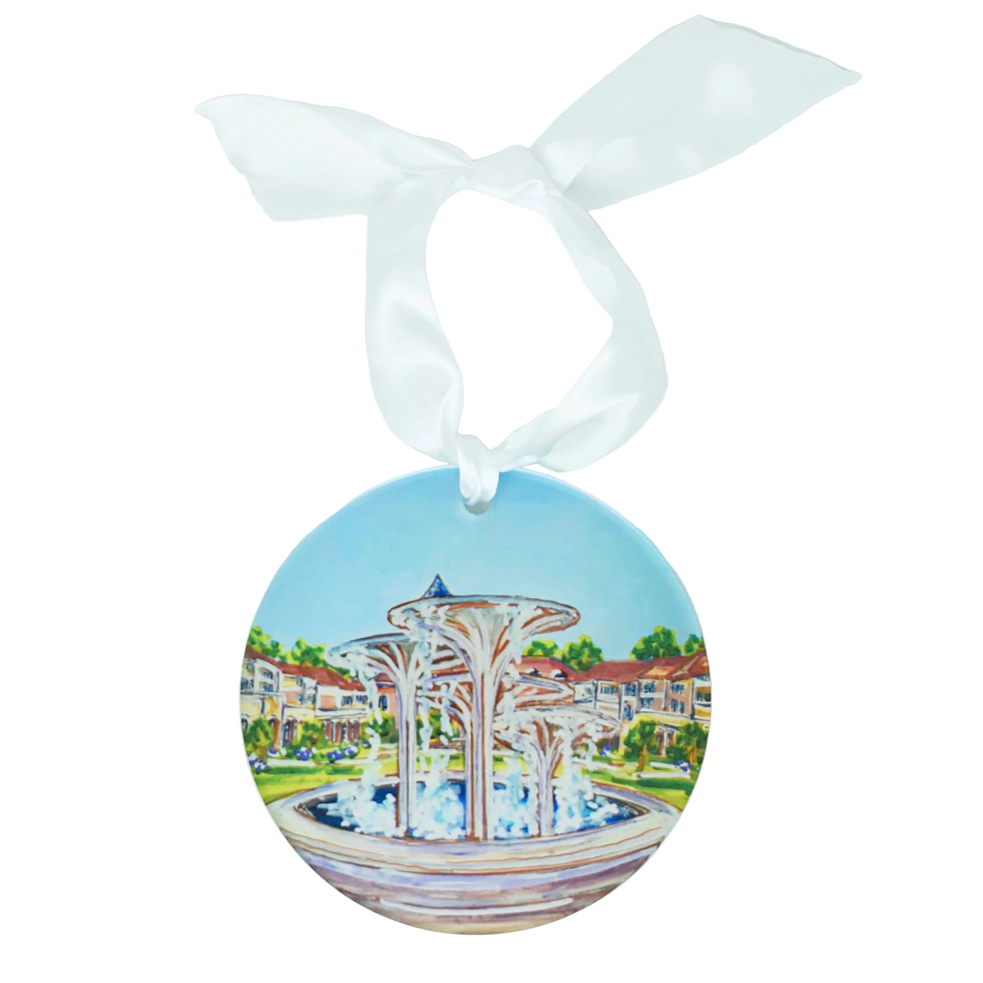 A round ceramic ornament featuring a painted illustration of a fountain in front of a resort-style building, hanging from a white satin ribbon bow on a white background.