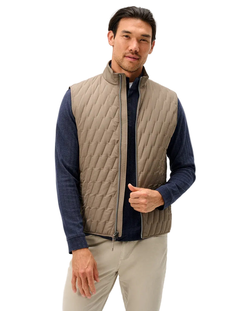 A man wearing a taupe quilted zip-front vest over a navy blue long-sleeve shirt and beige trousers, standing against a light gray studio background.