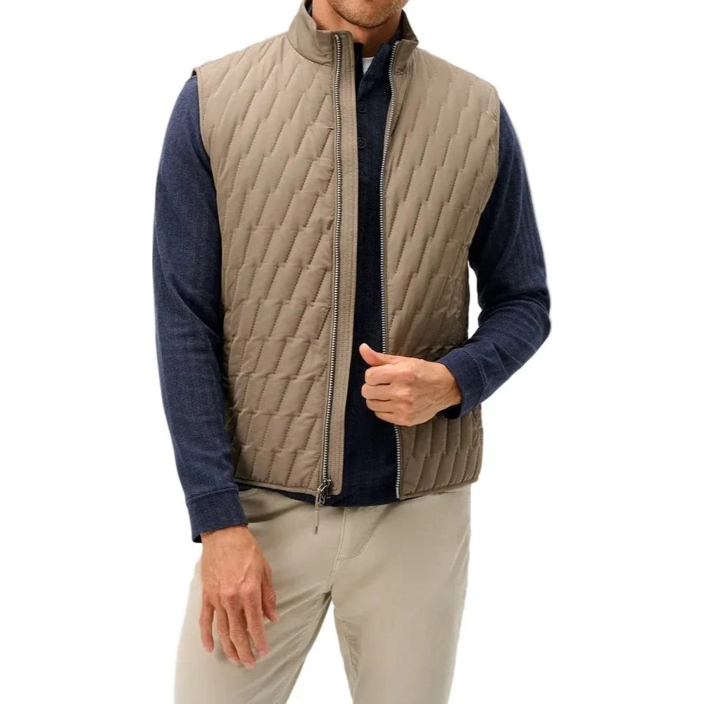 A man wearing a taupe quilted zip-front vest over a navy blue long-sleeve shirt and beige trousers, standing against a light gray studio background.