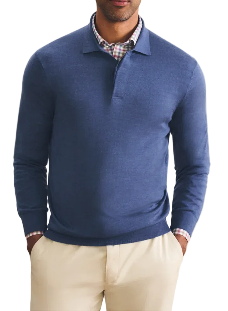 A man wearing a blue long-sleeve polo-neck sweater over a checkered collared shirt and cream trousers, standing against a warm beige textured background.