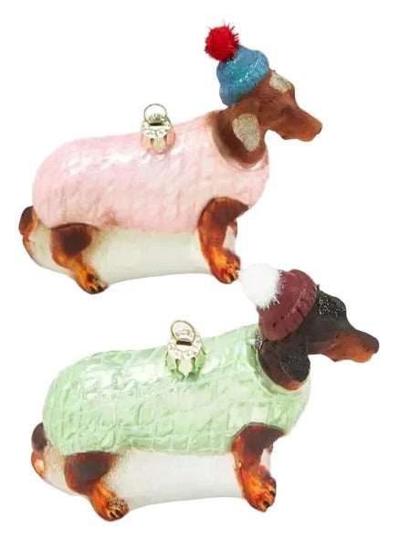 Two dachshund-shaped ornaments dressed in winter sweaters, one in pink with a blue hat and red pom-pom, and one in mint green with a burgundy hat and white pom-pom, on a light gray background.