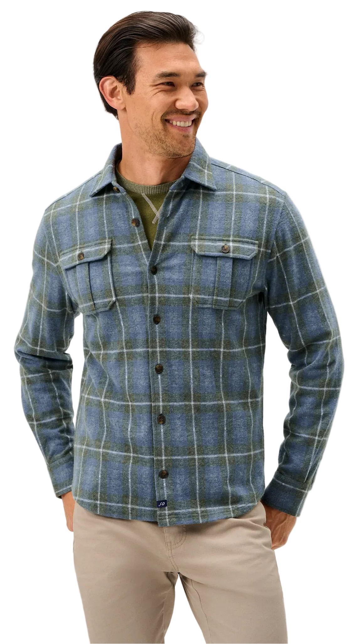A man wearing a blue and green plaid button-down flannel shirt over an olive green undershirt, paired with beige pants, against a light gray studio background.