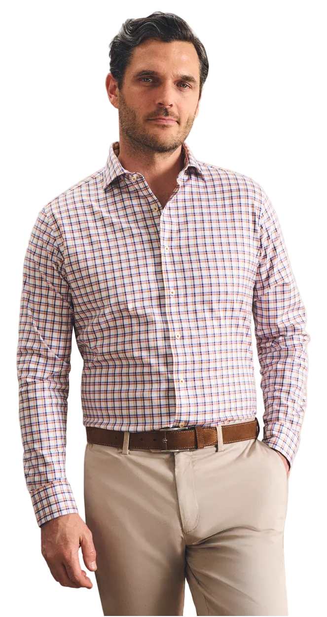 A man wearing a long-sleeve button-down shirt with a red, blue, and white gingham pattern, paired with beige trousers and a brown belt, standing against a textured beige wall.