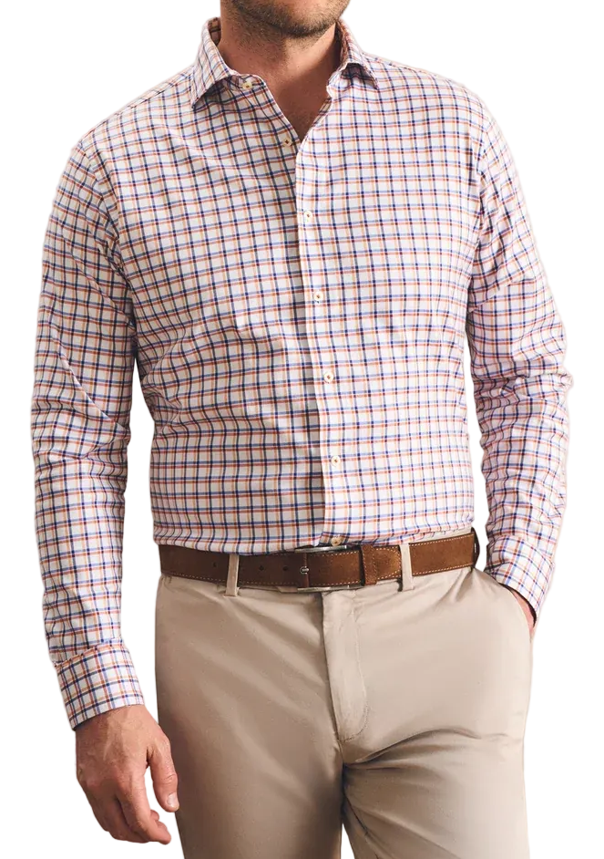 A man wearing a long-sleeve button-down shirt with a red, blue, and white gingham pattern, paired with beige trousers and a brown belt, standing against a textured beige wall.