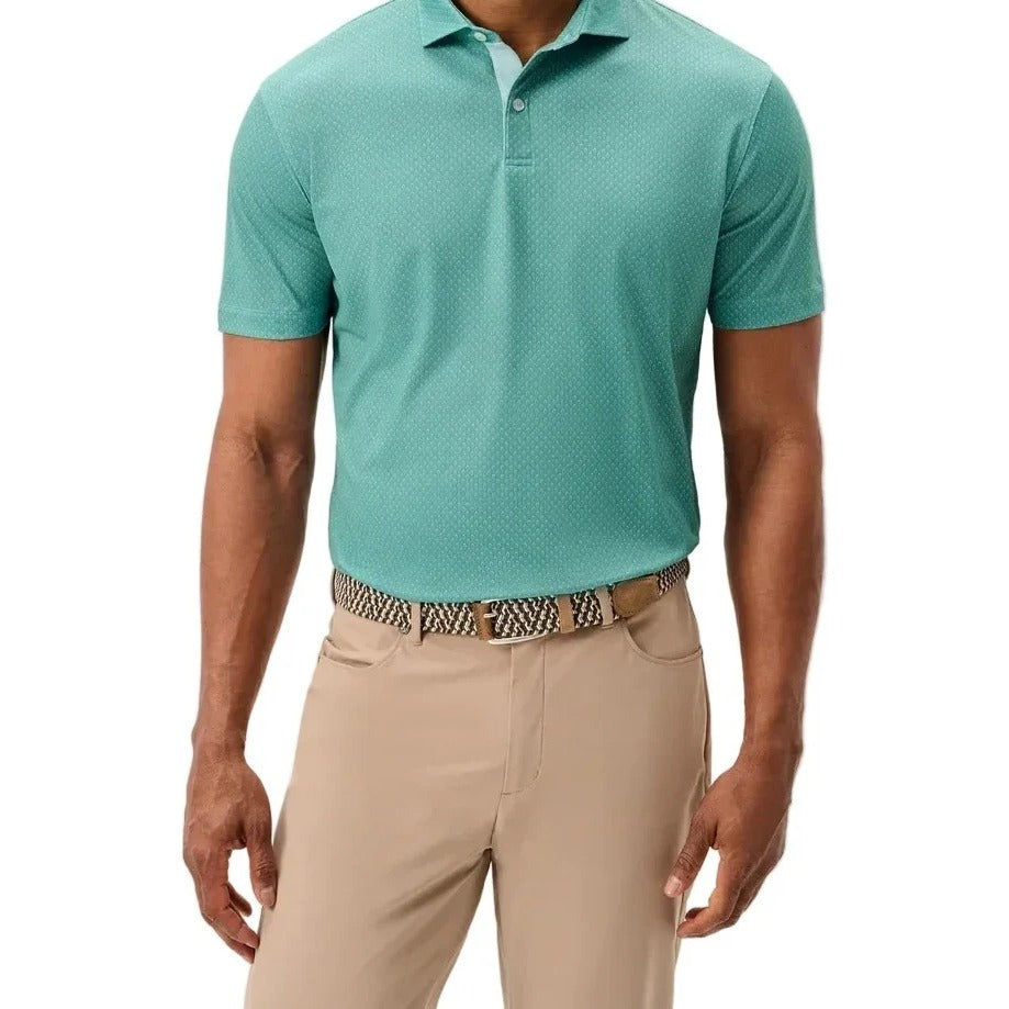 A man wearing a short-sleeve polo shirt in a muted teal-green color, paired with beige trousers and a patterned belt, standing against a light gray studio background.