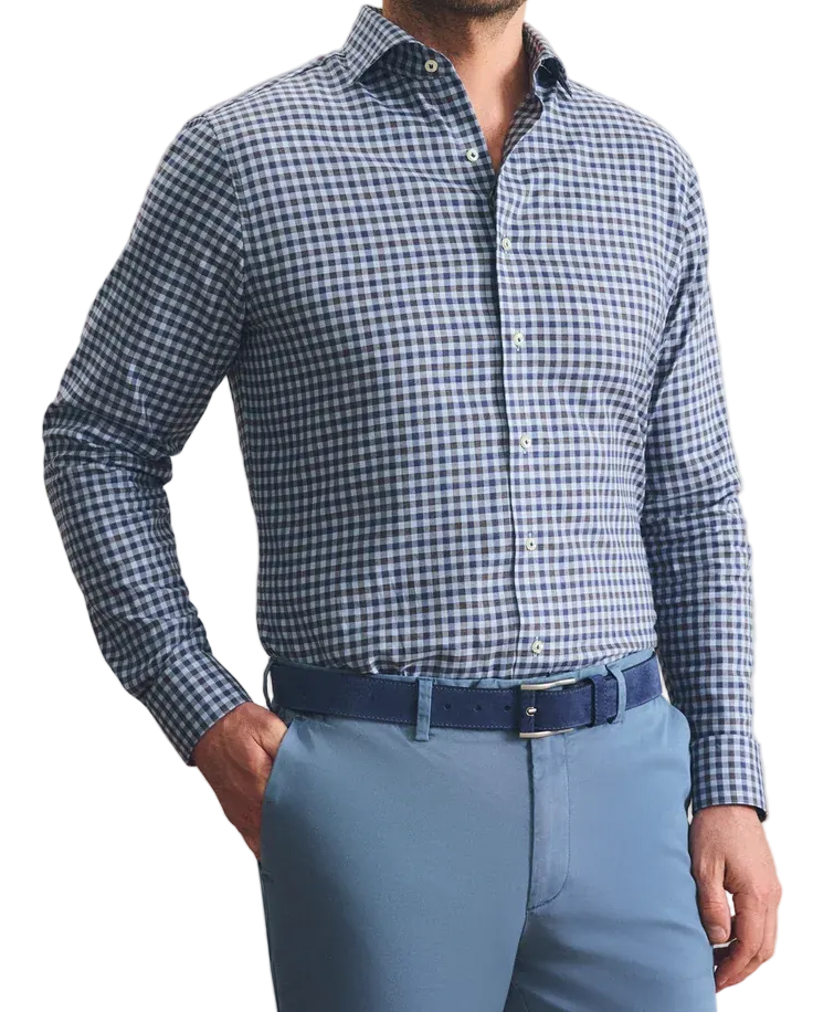 A man wearing a blue and white gingham long-sleeve dress shirt, paired with light blue trousers and a dark blue belt, standing against a textured beige wall.