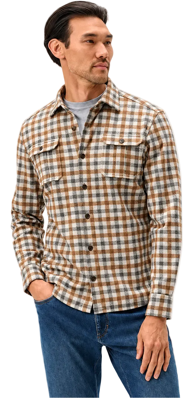 A man wearing a brown and beige plaid button-down flannel shirt over a gray t-shirt and blue jeans, standing against a light gray studio background.