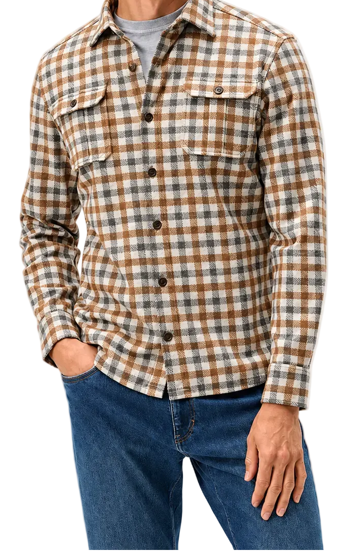 A man wearing a brown and beige plaid button-down flannel shirt over a gray t-shirt and blue jeans, standing against a light gray studio background.