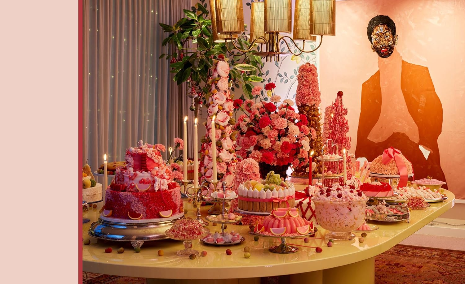 Decorative table with pink and red cake, flowers, and other treats against a wall with a painting.