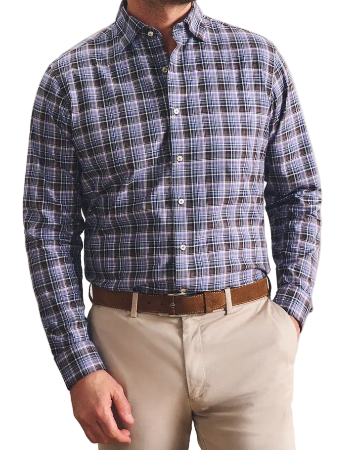 A man wearing a blue and gray plaid button-down shirt, beige trousers, and a brown belt, standing against a textured beige wall.