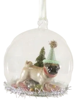 A clear glass Christmas ornament hanging from a white ribbon, featuring a small pug dog figurine wearing a green hat, set against a plain light background.