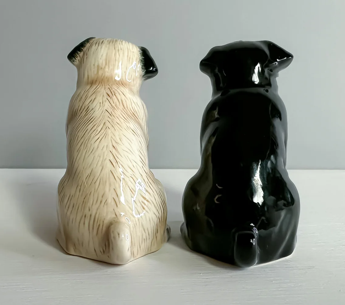 Two ceramic dog figurines, one black and one beige, on a white surface with a gray background.