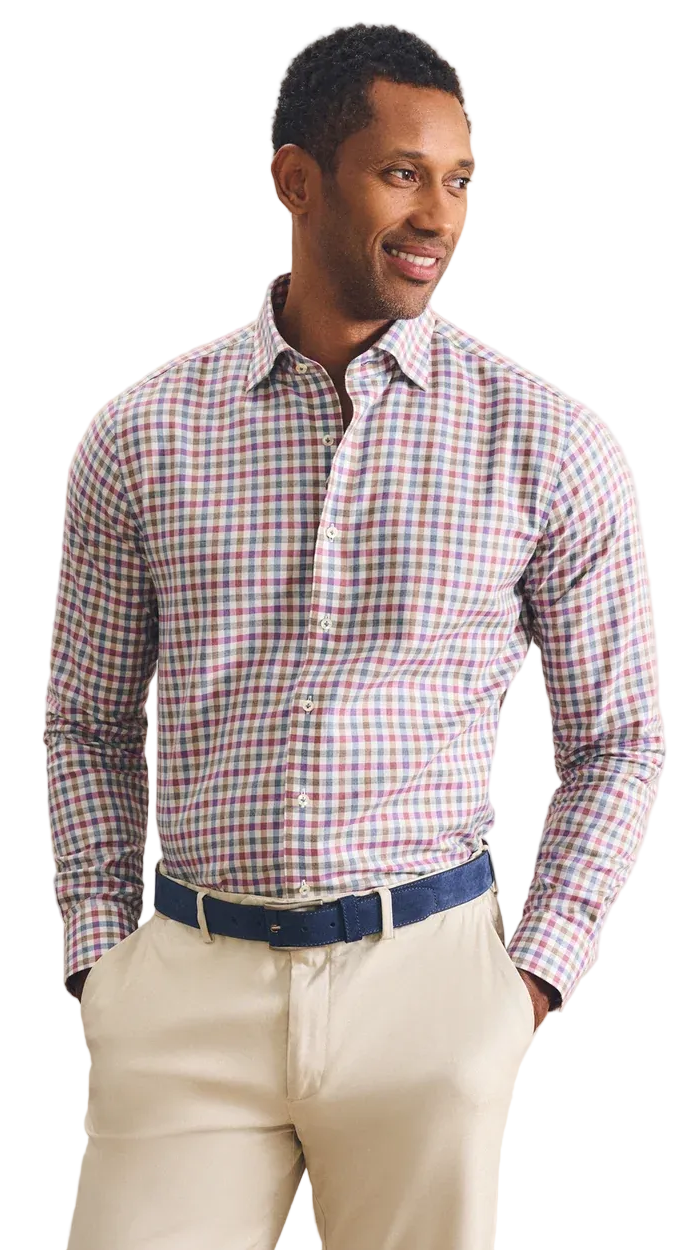 A man wearing a long-sleeve button-down shirt with a red, green, and white gingham pattern, paired with beige trousers and a navy blue belt, standing against a textured beige background.