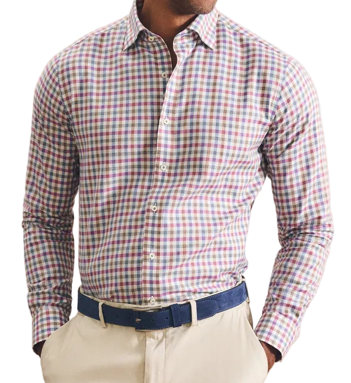 A man wearing a long-sleeve button-down shirt with a red, green, and white gingham pattern, paired with beige trousers and a navy blue belt, standing against a textured beige background.