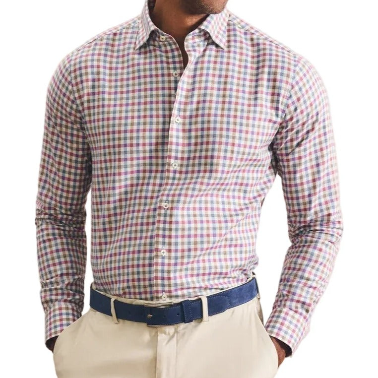 A man wearing a long-sleeve button-down shirt with a red, green, and white gingham pattern, paired with beige trousers and a navy blue belt, standing against a textured beige background.