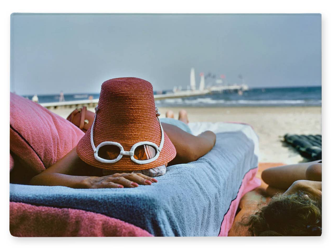 A rectangular wall art print showing a beach scene with a person lying on a lounge chair, wearing a brown hat and white-framed goggles. The print is displayed against a clean white background.