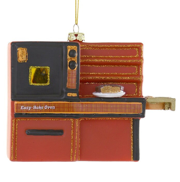 A Christmas ornament shaped like a retro easy-bake oven, featuring red and orange panels with gold glitter trim, a black front door, and a small cake on a saucer inside, hanging from a gold string against a white background.