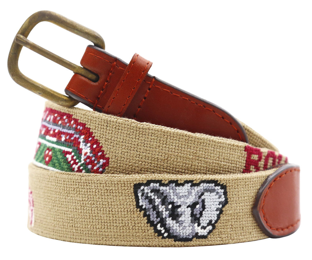 A woven canvas belt in tan with a brown leather buckle and keeper, featuring embroidered red, green, white, and gray details including a ram's head motif.