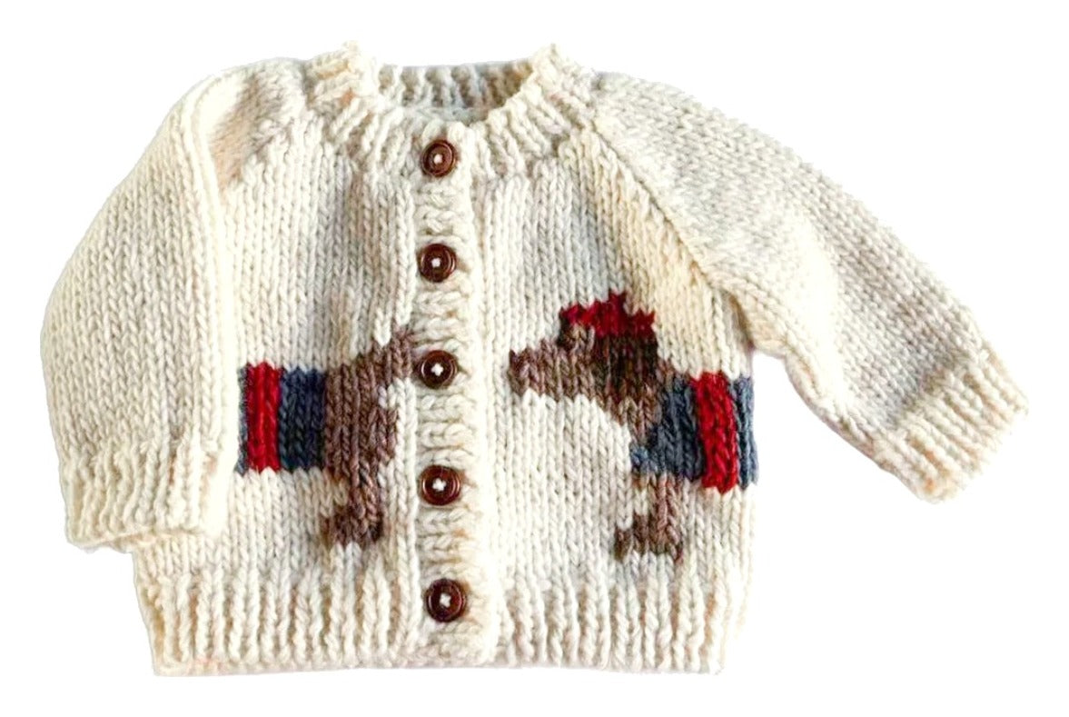 A cream-colored knit baby cardigan sweater featuring a knitted design of two horses facing each other, with a red and blue striped band between them. The cardigan has brown buttons down the center front.