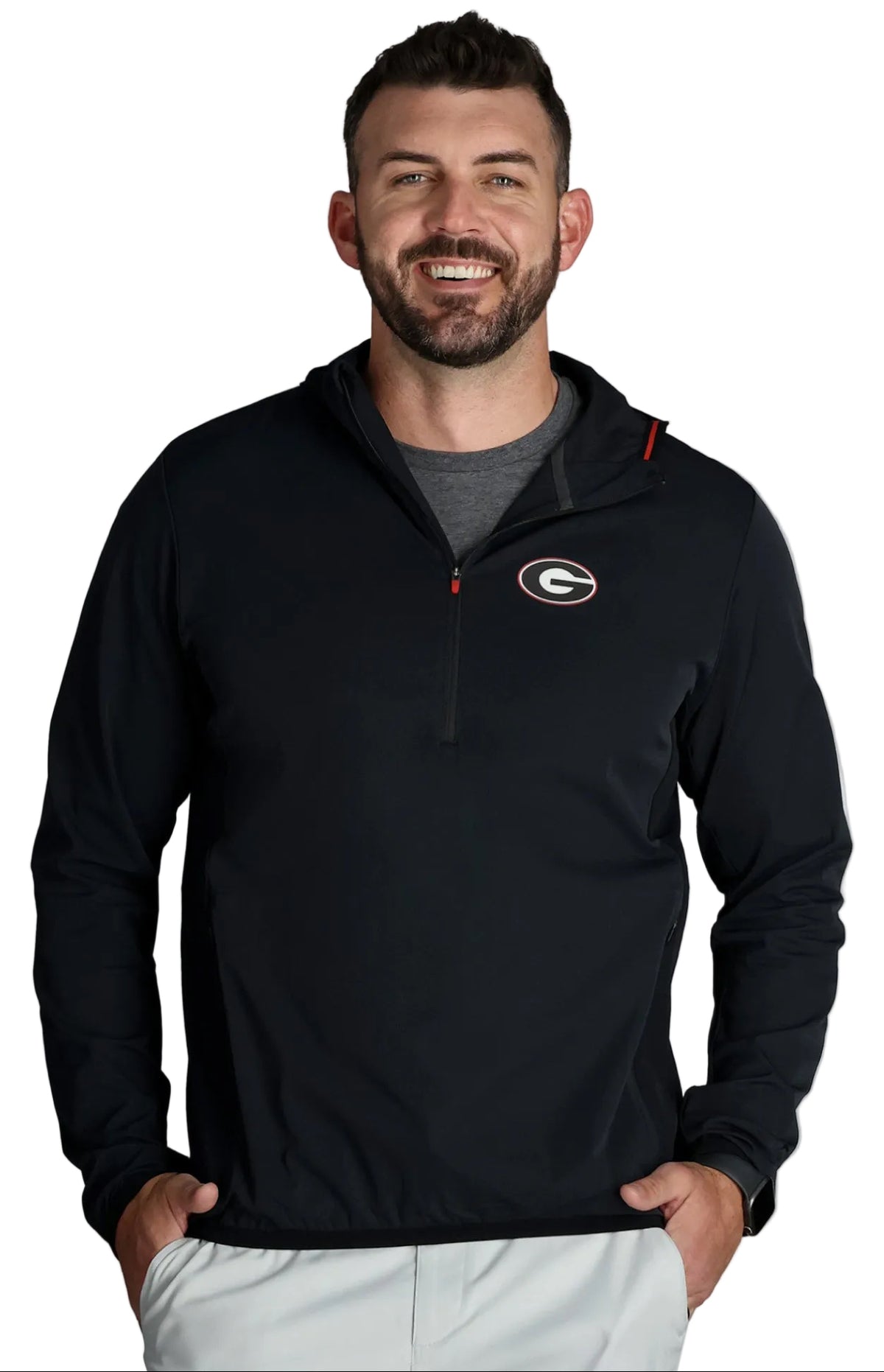 A smiling man wearing a black half-zip pullover jacket with a Georgia Bulldogs logo on the left chest, paired with a gray t-shirt and light gray pants, against a light gray studio background.