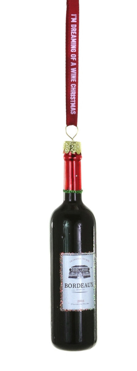 A Christmas ornament shaped like a Bordeaux wine bottle, featuring a dark glass body, a red neck with a gold ornament loop, and a white label. A red hanging ribbon at the top reads, 'I'M DRINKING OF A WINE CHRISTMAS'.