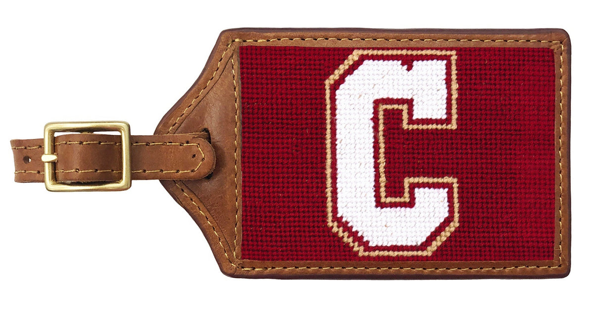 A rectangular leather luggage tag with a brown leather strap and buckle, featuring a red needlepoint canvas face with a large white letter C outlined in gold.