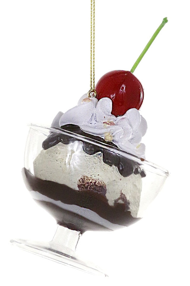 A clear glass dessert-shaped ornament hanging from a gold string, decorated with white whipped cream, dark chocolate sauce, and a red cherry on a green stem, against a plain white background.
