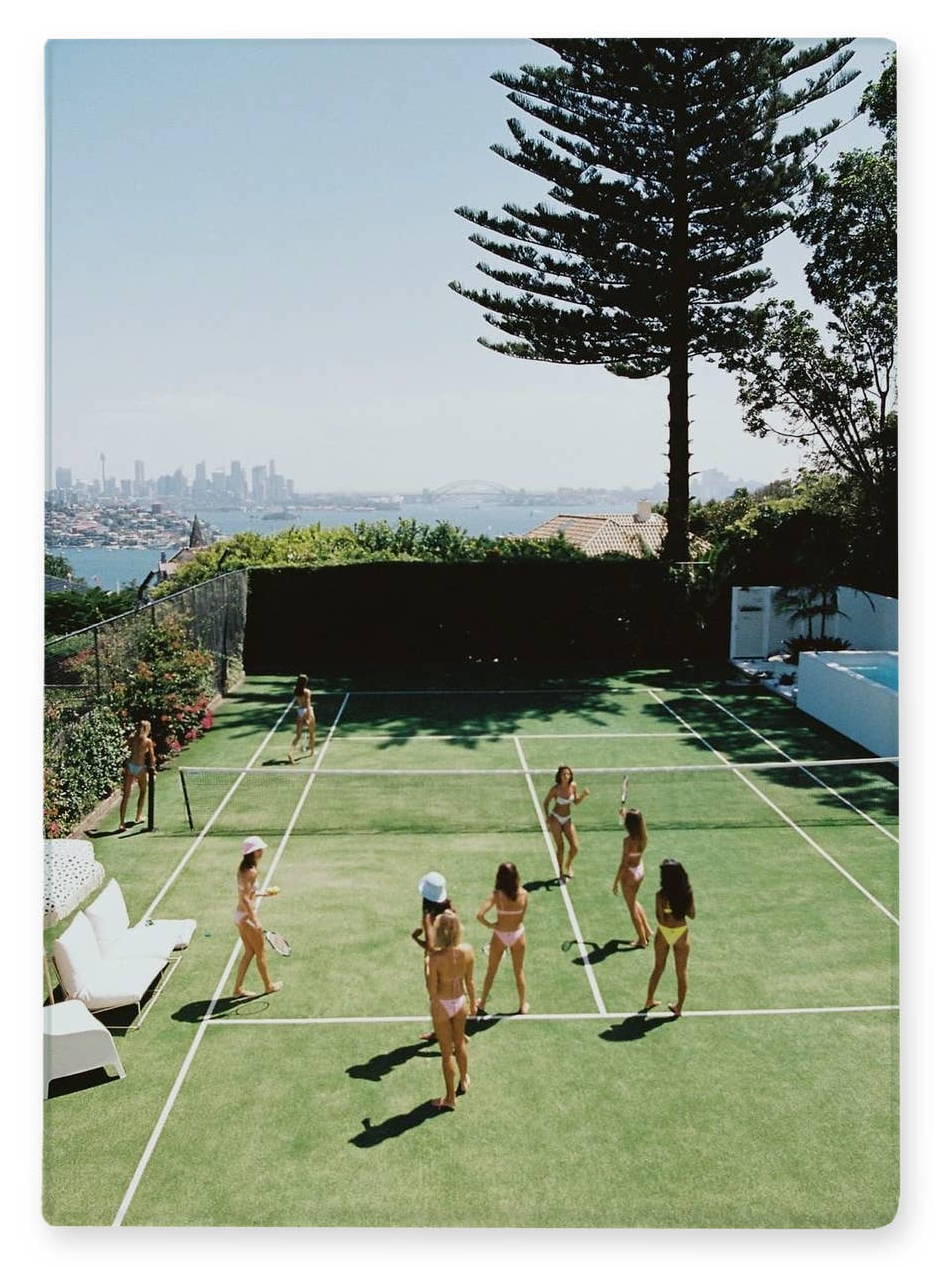 A vertical photographic print showing a sunny outdoor tennis court scene with several women playing on green grass, surrounded by a tall pine tree, lounge chairs, and a distant city skyline over water. The print is displayed against a clean white background.