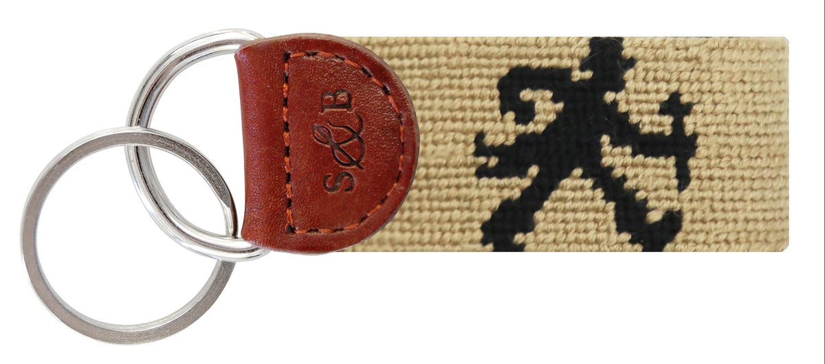 A woven canvas keychain with a black embroidered figure motif on a beige background, attached to a brown leather tab embossed with 'S&P' and two silver metal key rings.
