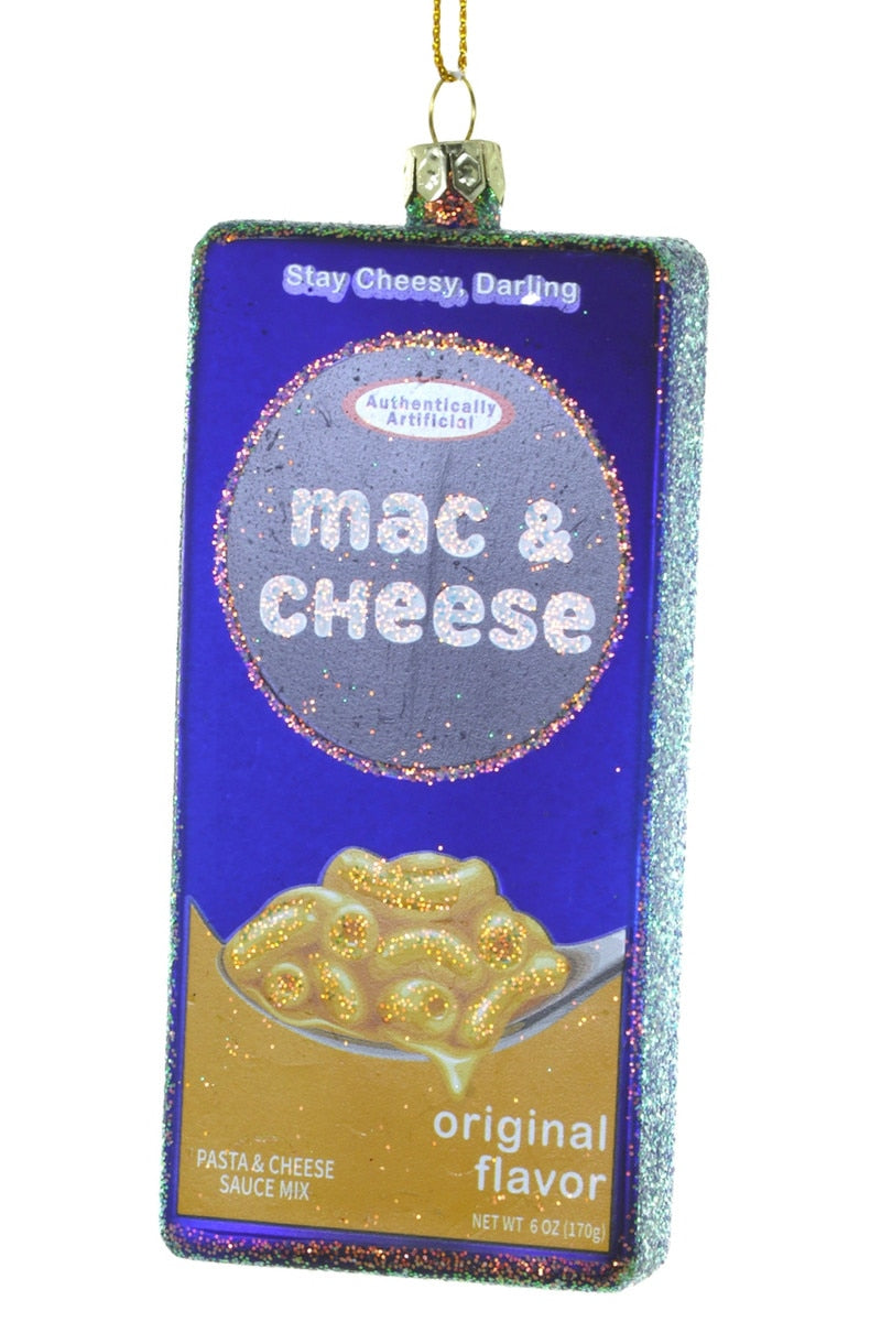 A rectangular Christmas ornament shaped like a box of mac and cheese sauce mix, featuring a purple and gold design with the words 'Stay Cheesy, Darling' and 'mac & cheese'.